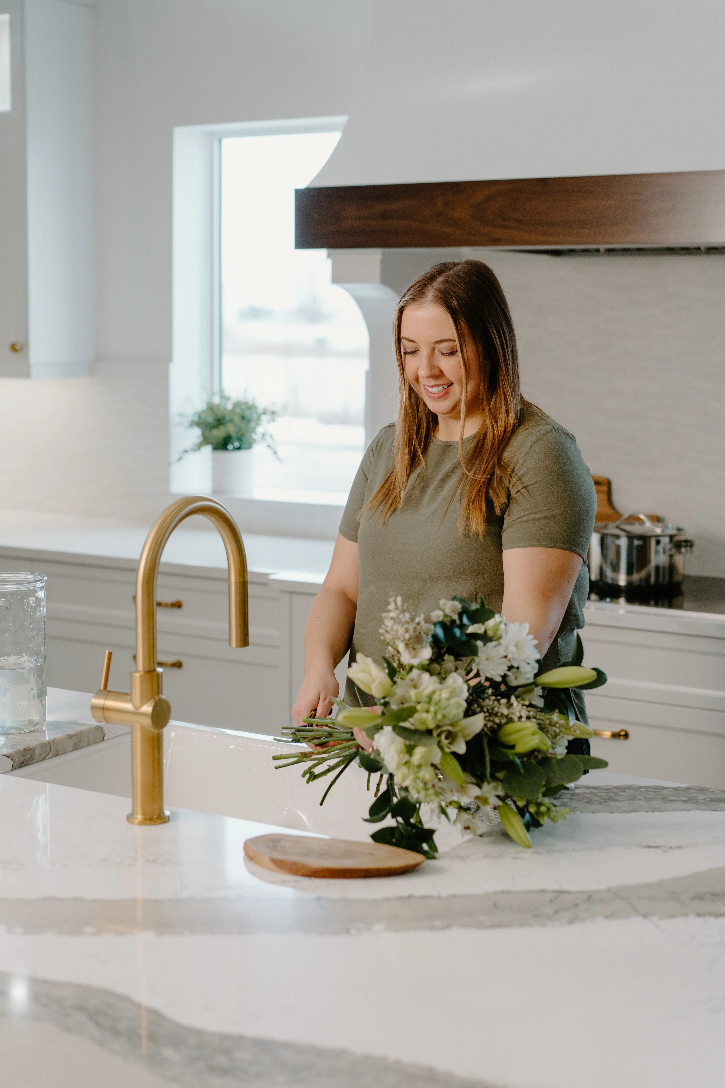 Woman preparing flowers in a modern kitchen with a white marble countertop and brass faucet.