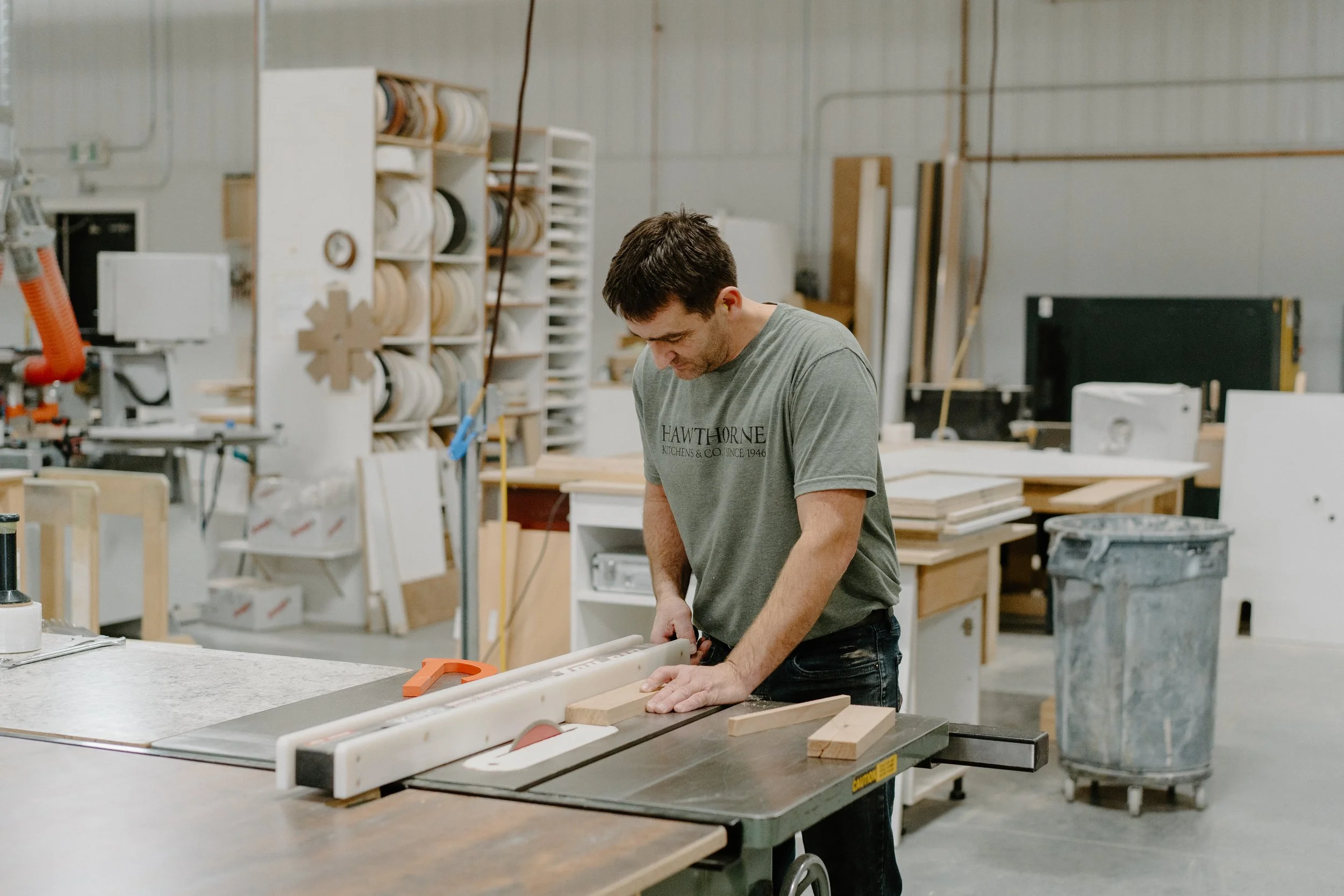 A man working in a woodworking shop, sawing a piece of wood on a table saw.