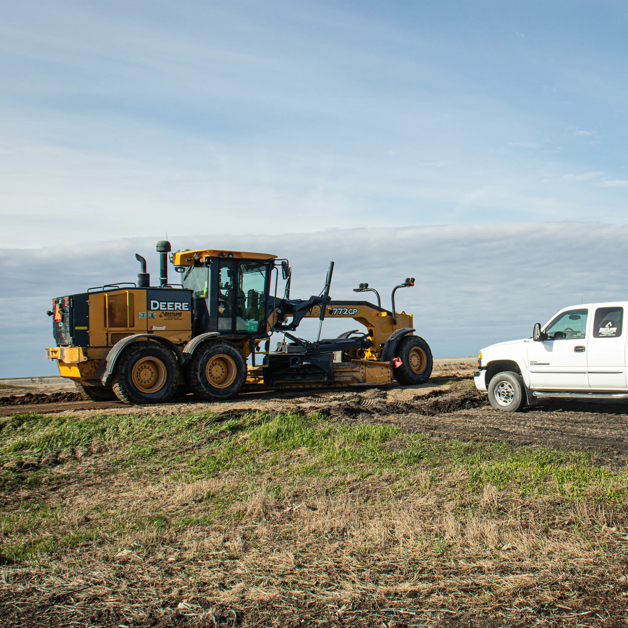 John Deere 772GP grader working alongside a white pickup truck on a rural road building project.