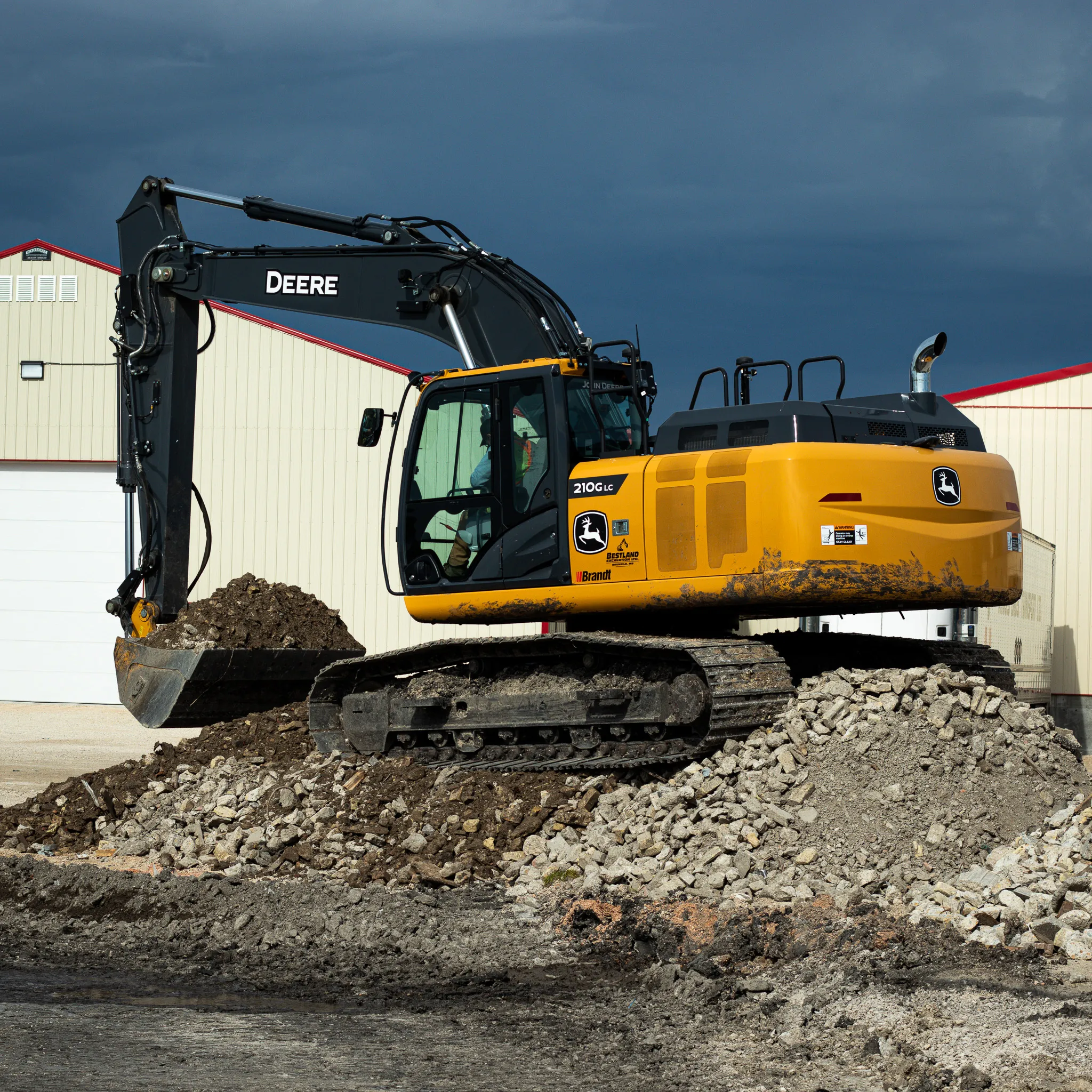 John Deere 210G LC excavator perched on a pile of rubble, scooping soil in front of a large beige commercial shop.
