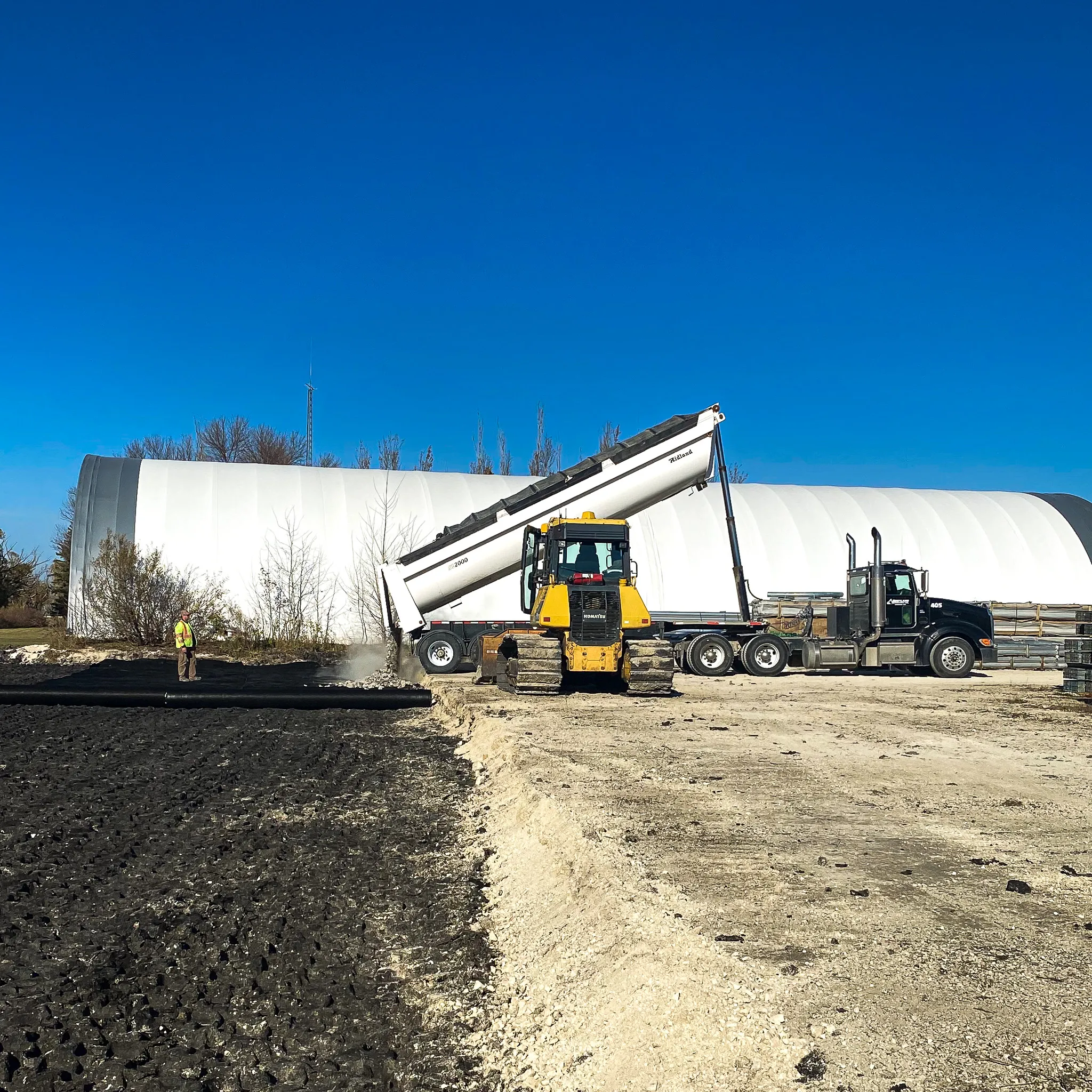 Dump truck unloading materials while a bulldozer levels ground near a large white agricultural storage building.
