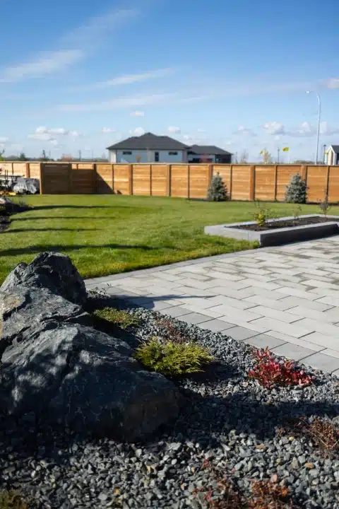 Backyard with a grey paver patio, large boulder surrounded by small shrubs, &amp; wooden privacy fence.