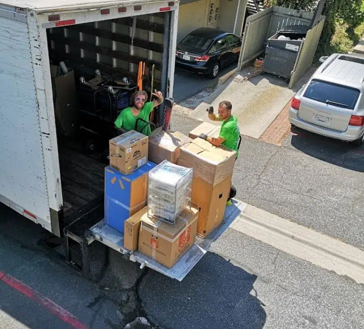Two movers in green shirts load boxes onto a moving truck parked in a residential street. The truck