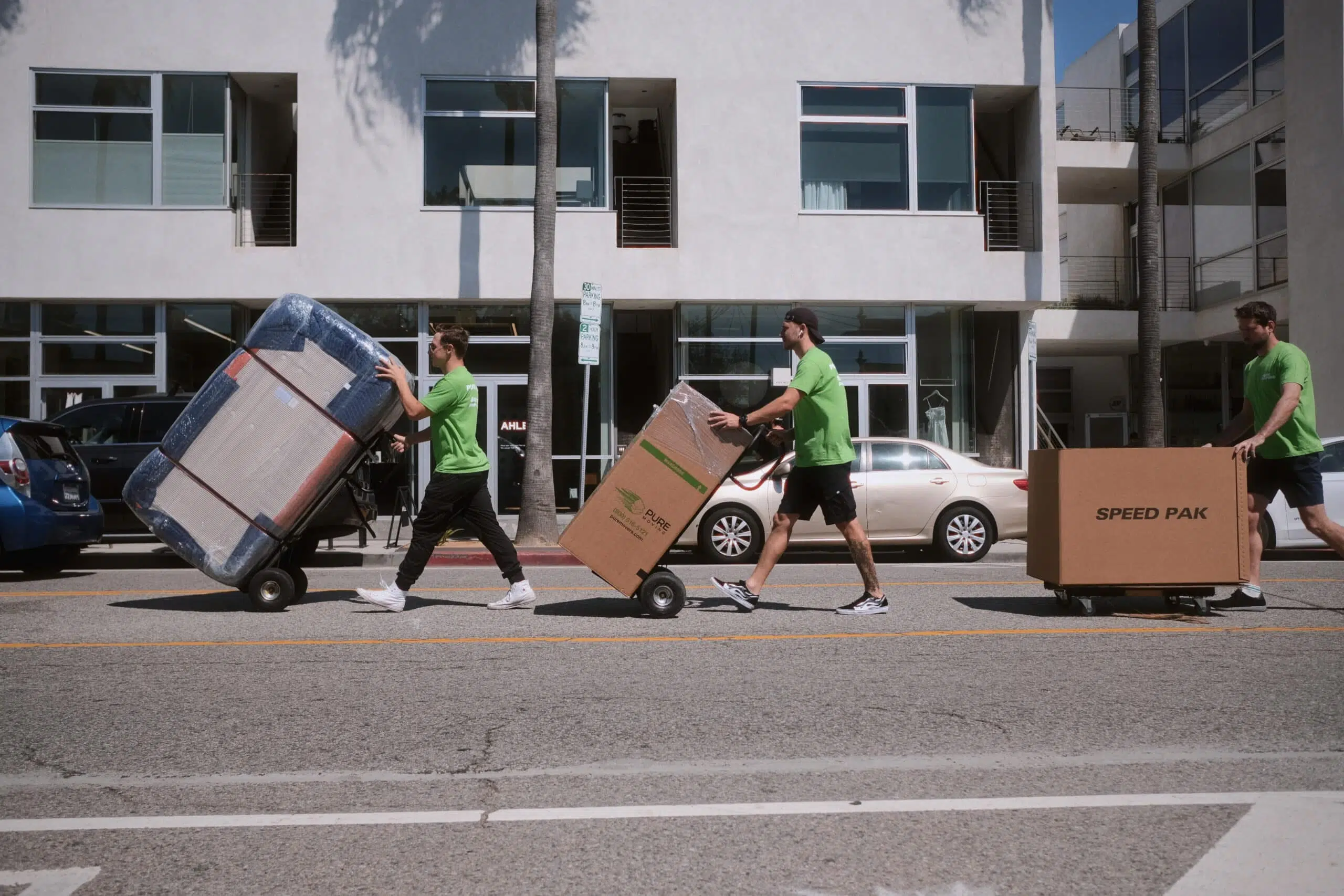 Three movers in green shirts, offering top-notch Moving and Packing Services, push items on dollies across a street in f