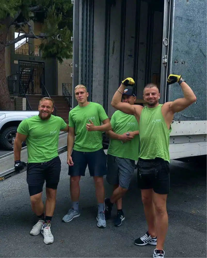 Four men in green shirts and shorts from Bay Area Movers are standing near an open truck. Two of them are posing with th