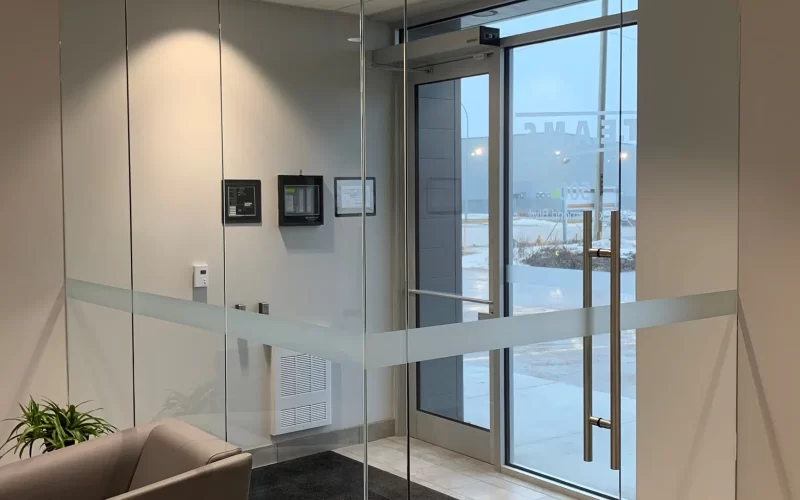 A modern office entrance with security glass doors and neutral-colored walls greets visitors. A small plant sits on a ta