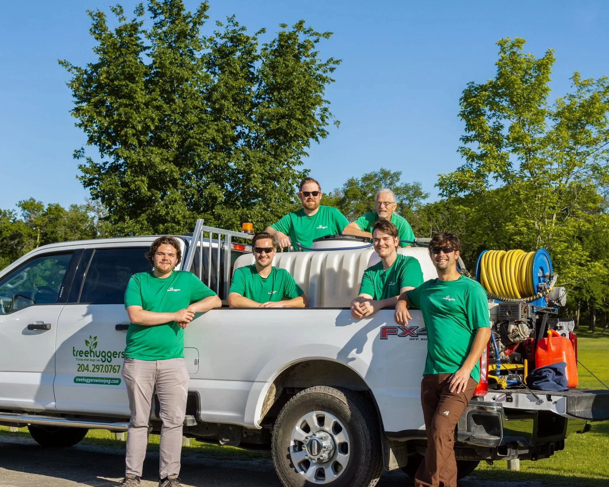 A group of six men in green shirts standing in front of a white truck with equipment mounted on it, including hoses, too